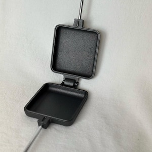 May include: A black cast iron s'mores maker with a silver handle. The maker is open, revealing two square cooking surfaces.