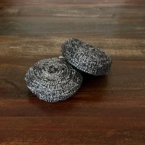 May include: Two silver-coloured scouring pads, made of tightly woven metal, are stacked on top of each other on a wooden surface.