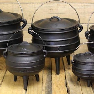 May include: A set of five black cast iron pots with lids, each featuring three legs and a wire handle. The pots vary in size and are arranged in a group, showcasing a rounded body design with horizontal ridges on the exterior surface.