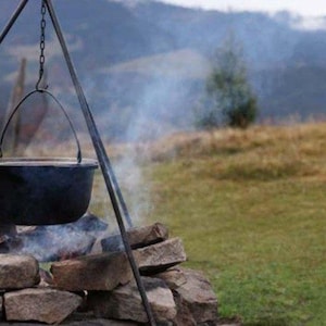 May include: A black metal cauldron hangs from a tripod stand over a stone fire pit. The pot is suspended by a metal chain and handle, with visible smoke rising from the fire below, set against a backdrop of grassy hills and mountains.
