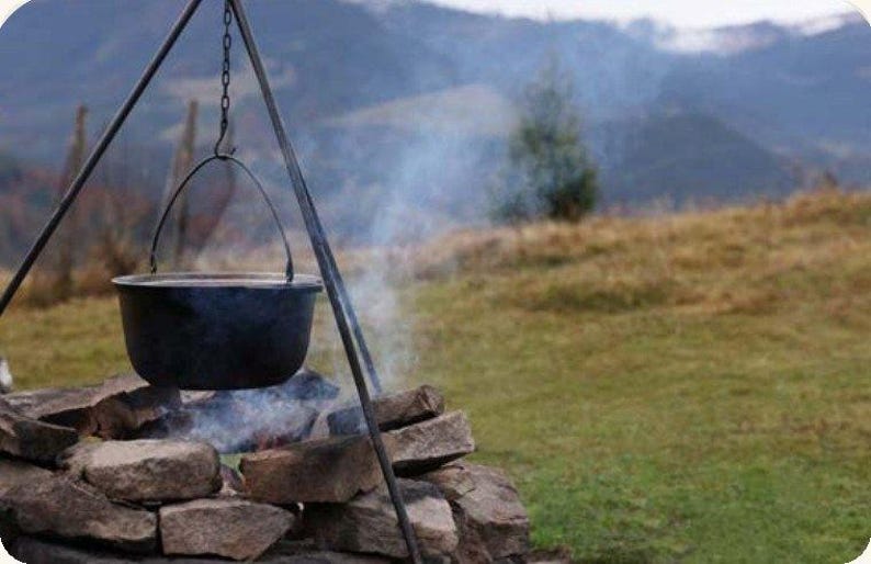 May include: A black metal cauldron hangs from a tripod stand over a stone fire pit. The pot is suspended by a metal chain and handle, with visible smoke rising from the fire below, set against a backdrop of grassy hills and mountains.
