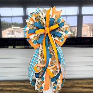 May include: A decorative bow featuring orange, teal, and white ribbons. The bow has a pumpkin and leopard print pattern, with solid teal and orange ribbons. The bow is mounted on a gray wooden base, perfect for fall decor.