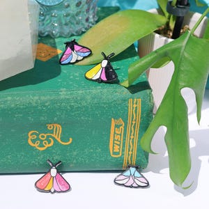 May include: Four enamel pins featuring colorful butterfly designs. The pins are arranged on a green book with the word "WISE" printed on the spine. The book is sitting on a white surface.