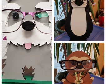 May include: Three wooden animal eyeglass holders. A gray schnauzer, a black and white penguin, and a brown sloth. Each animal is holding a pair of eyeglasses.