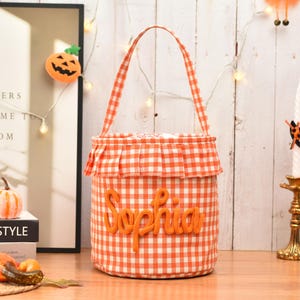 May include: A cylindrical fabric bucket bag featuring an orange and white gingham check pattern. The bag has a single handle, a ruffled trim around the top edge, and the name Sophia embroidered in orange yarn lettering on the front center.
