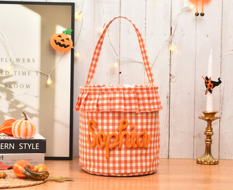 May include: A cylindrical fabric bucket bag featuring an orange and white gingham check pattern. The bag has a single handle, a ruffled trim around the top edge, and the name Sophia embroidered in orange yarn lettering on the front center.