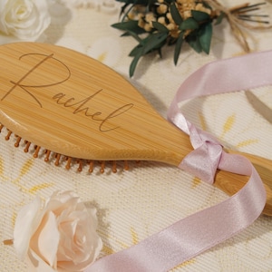 May include: A wooden hairbrush engraved with the name "Rachel". The brush is light brown and tied with a light pink ribbon. The bristles are a darker brown. The background features decorative flowers and greenery. The brush is approximately 20 cm long.