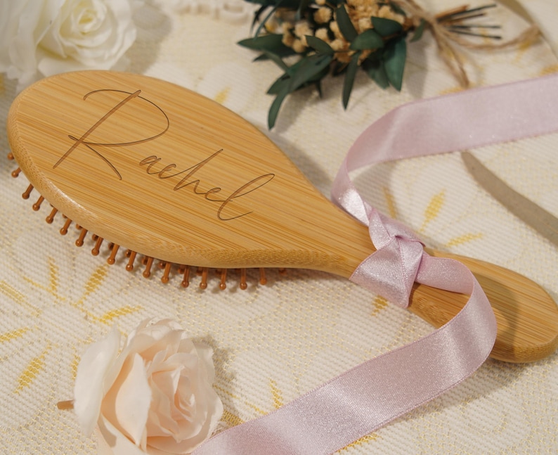 May include: A wooden hairbrush engraved with the name "Rachel". The brush is light brown and tied with a light pink ribbon. The bristles are a darker brown. The background features decorative flowers and greenery. The brush is approximately 20 cm long.