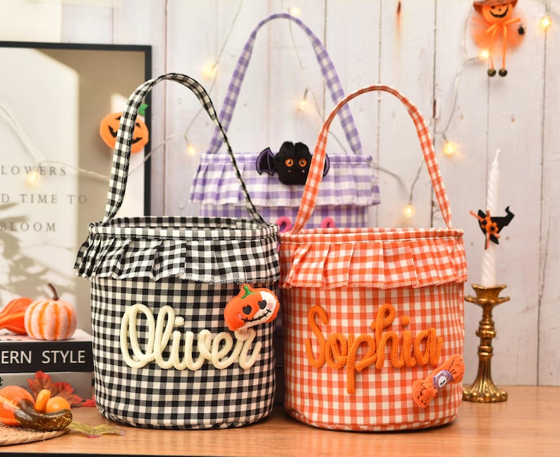 May include: Three cylindrical fabric baskets with handles and ruffled tops. One is black and white gingham with the name Oliver, one is orange and white gingham with the name Sophia, and one is purple and white gingham with a bat applique.