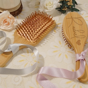 May include: Two wooden hairbrushes with light brown bristles. One is rectangular, the other oval-shaped and engraved with the name "Rachel" and a heart. Both brushes are tied with satin ribbons, one silver and one pink.