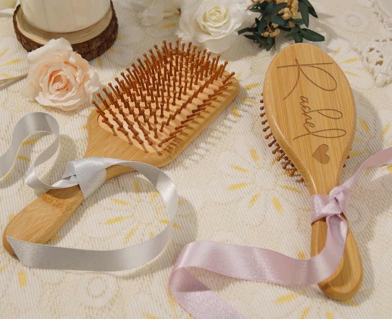 May include: Two wooden hairbrushes with light brown bristles. One is rectangular, the other oval-shaped and engraved with the name "Rachel" and a heart. Both brushes are tied with satin ribbons, one silver and one pink.