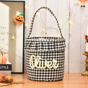 May include: A cylindrical fabric basket featuring a black and white gingham check pattern, a ruffled top edge, and a single handle. The name Oliver is written in large, cream-colored, raised script lettering across the front of the basket.