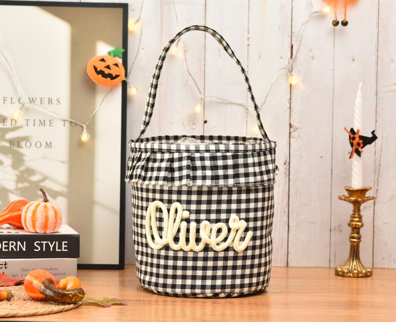 May include: A cylindrical fabric basket featuring a black and white gingham check pattern, a ruffled top edge, and a single handle. The name Oliver is written in large, cream-colored, raised script lettering across the front of the basket.