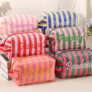 Puede incluir: Varias bolsas de cosméticos a rayas en varios colores, incluyendo rosa, rojo, naranja, verde y azul. Cada bolsa tiene un nombre bordado en un color contrastante. Las bolsas están dispuestas sobre una superficie blanca.
