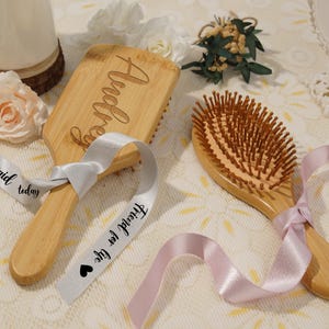 May include: Two wooden hairbrushes with light brown bristles and handles. One brush is engraved with the name "Audrey". Both brushes are adorned with satin ribbons. One ribbon reads "Bridesmaid today" and "Friend for life".