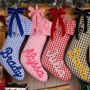 May include: Four Christmas stockings in blue, pink, red, and green gingham patterns. Each stocking features a ruffled cuff, a velvet bow, and a personalised name embroidered in a cursive rope-style font: Brady, Alyssa, Julia, and Pamela.