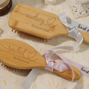 May include: Two wooden hairbrushes, engraved with names and the word "Bridesmaid". Each brush has a ribbon attached, one grey and one pink, with the text "Friend for life". The brushes are a light brown colour.