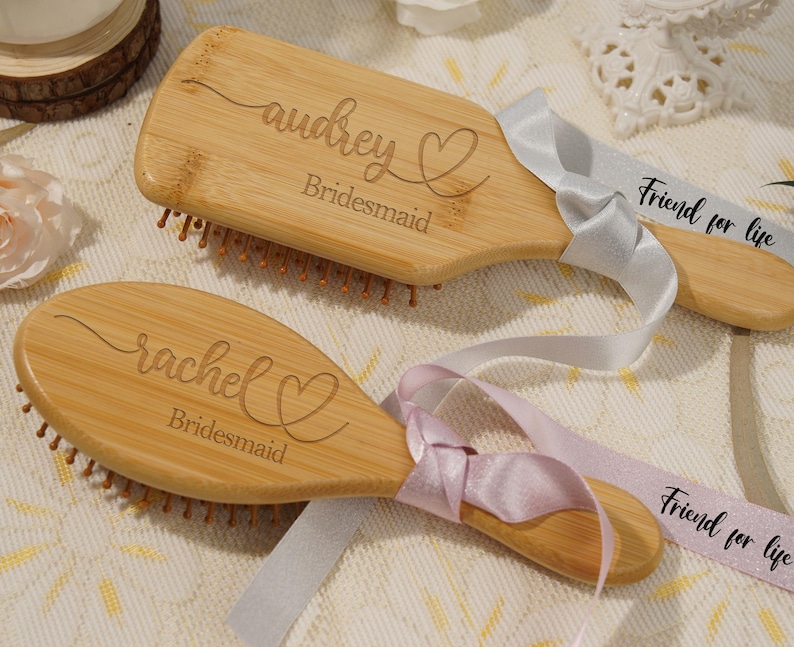 May include: Two wooden hairbrushes, engraved with names and the word "Bridesmaid". Each brush has a ribbon attached, one grey and one pink, with the text "Friend for life". The brushes are a light brown colour.