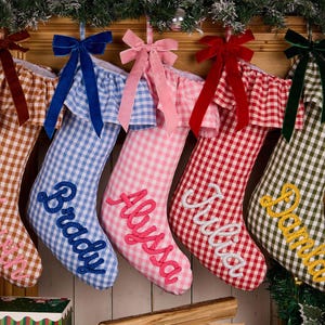 May include: Five fabric Christmas stockings with a gingham pattern in brown, blue, pink, red, and green. Each stocking features a ruffled cuff, a velvet bow, and a name embroidered in cursive: Henry, Brady, Alyssa, Julia, and Damla.