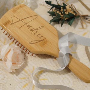 May include: A wooden hairbrush with the name "Audrey" engraved on the handle. The brush has brown bristles and a light wood finish. A silver ribbon and a light pink rose are next to the brush.