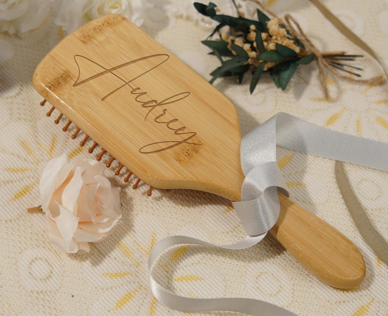 May include: A wooden hairbrush with the name "Audrey" engraved on the handle. The brush has brown bristles and a light wood finish. A silver ribbon and a light pink rose are next to the brush.