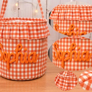 May include: A cylindrical fabric basket featuring an orange and white gingham pattern. The basket has a ruffled top edge and a handle. The name Sophia is embroidered in orange yarn on the front. Circular insets show the ruffle and fabric detail.