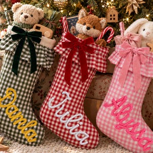 Puede incluir: Tres calcetines navideños de cuadros vichy en verde, rojo y rosa, cada uno con un puño con volantes, un lazo de terciopelo y un peluche. Los calcetines están bordados con los nombres Damla en amarillo, Julia en blanco y Allyssa en rosa.