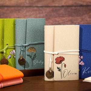 May include: A collection of fabric-covered journals in lime green, teal, beige, and blue. Each journal features a floral illustration, a name in script, and a cord closure with a small round metal clock charm attached to the front cover.