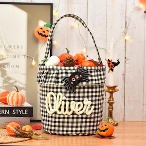 May include: A black and white gingham fabric bucket with a ruffled top edge and a single handle. The front features a black spider appliqu&eacute; with orange eyes and the name Oliver in white script lettering. The bucket is filled with small pumpkins.