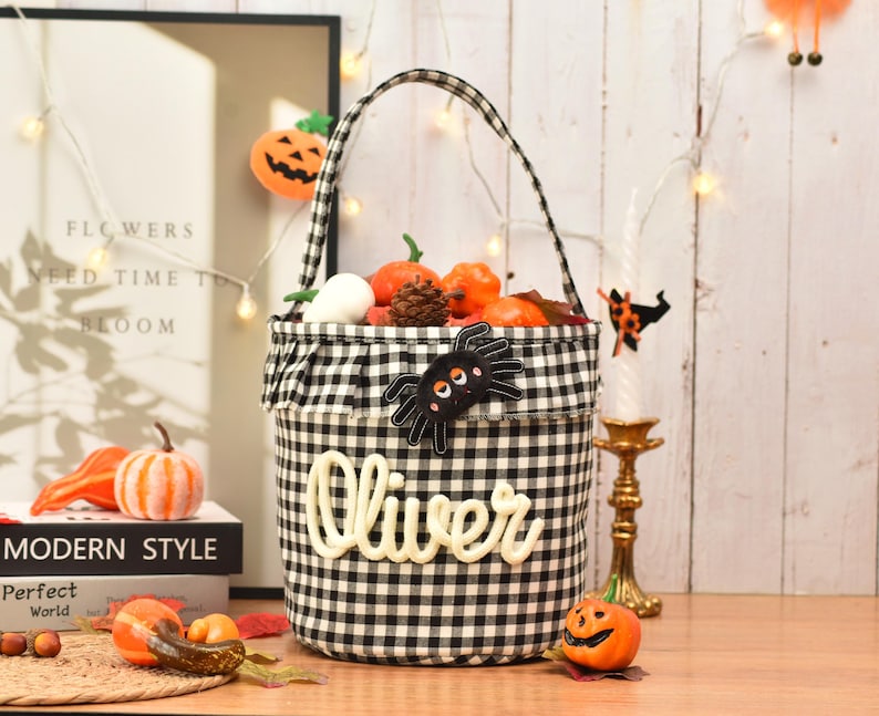 May include: A black and white gingham fabric bucket with a ruffled top edge and a single handle. The front features a black spider appliqu&eacute; with orange eyes and the name Oliver in white script lettering. The bucket is filled with small pumpkins.