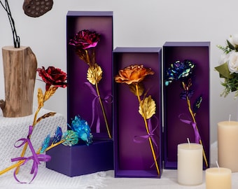 May include: Metallic roses in various colors, including red, purple, and blue, with gold-colored stems and leaves. Each rose is presented in a purple box and tied with a purple ribbon. The arrangement includes candles and a wooden vase.