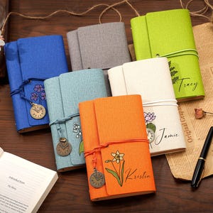 May include: A collection of six fabric-covered journals in blue, gray, light blue, orange, cream, and lime green. Each notebook features a wrap-around cord, a hanging clock charm, and a floral illustration with a personalized name printed on the cover.