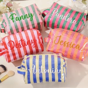 Peut inclure: Cinq trousses de maquillage personnalisées à rayures verticales de différentes couleurs : rose et vert, rose et rouge, rose et orange, et jaune et bleu. Chaque trousse porte un nom différent : Fanny, Pandora, Destiny, Jessica et Antonia.