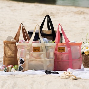 Könnte beinhalten: Mehrere Strandtaschen aus Mesh in verschiedenen Farben, darunter Beige, Schwarz und Rosa, werden an einem Sandstrand gezeigt. Jede Tasche hat einen kontrastierenden Griff und ein Namensschild. Sonnenbrillen, Sandalen und ein Blumenkorb sind ebenfalls vorhanden.