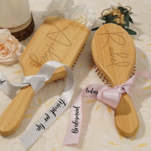 May include: Two wooden hairbrushes with engraved names, tied with ribbons. One brush is rectangular, the other oval. Ribbons say "Friend for life" and "Bridesmaid". The brushes are light brown with dark brown bristles, and the ribbons are white and pink.