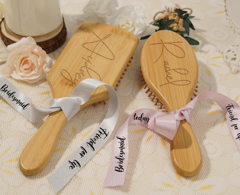 May include: Two wooden hairbrushes with engraved names, tied with ribbons. One brush is rectangular, the other oval. Ribbons say "Friend for life" and "Bridesmaid". The brushes are light brown with dark brown bristles, and the ribbons are white and pink.