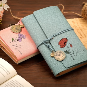 May include: Two fabric-covered journals, one light blue and one pink, each featuring a floral illustration, a name printed in script, and a dangling antique-style clock charm. The blue journal is wrapped with a matching cord closure.