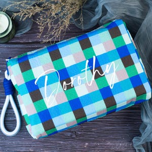 May include: A blue, green, and brown plaid makeup bag with a white zipper and a white rope handle. The bag has the name "Dorothy" written in white on the front.