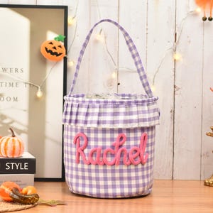 May include: A cylindrical fabric basket featuring a purple and white gingham pattern with a ruffled top edge and a single handle. The name Rachel is embroidered in pink yarn across the front of the basket, which has a structured, upright design.