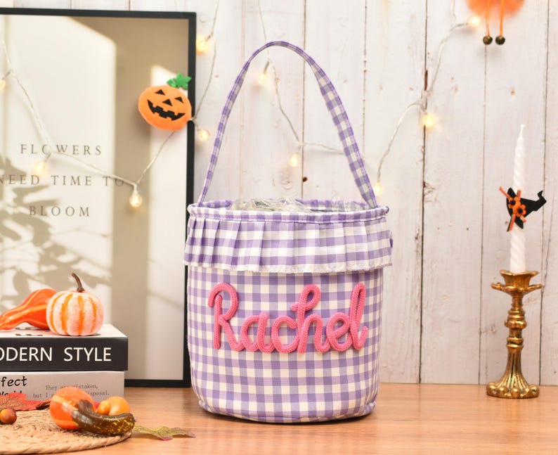 May include: A cylindrical fabric basket featuring a purple and white gingham pattern with a ruffled top edge and a single handle. The name Rachel is embroidered in pink yarn across the front of the basket, which has a structured, upright design.