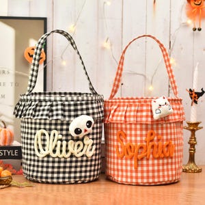 May include: Two cylindrical fabric baskets with handles, one in black and white gingham and one in orange and white gingham. Each basket features a ruffled top edge, a plush ghost or cat accent, and a name embroidered in cursive script.