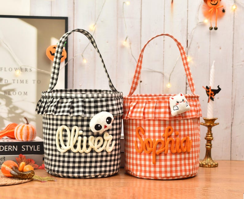 May include: Two cylindrical fabric baskets with handles, one in black and white gingham and one in orange and white gingham. Each basket features a ruffled top edge, a plush ghost or cat accent, and a name embroidered in cursive script.