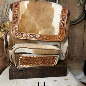 May include: A backpack crafted from tan and white cowhide featuring a large top flap and a smaller front pocket, both accented with intricate brown leather tooling in a floral pattern. The bag includes dark brown zippers and a top handle.