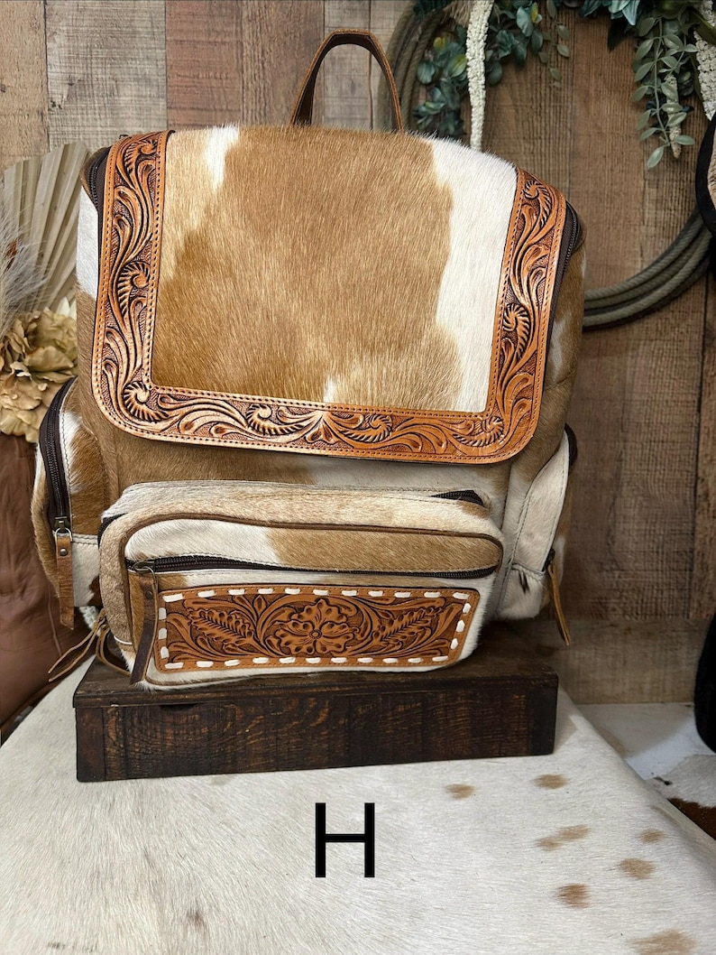 May include: A backpack crafted from tan and white cowhide featuring a large top flap and a smaller front pocket, both accented with intricate brown leather tooling in a floral pattern. The bag includes dark brown zippers and a top handle.