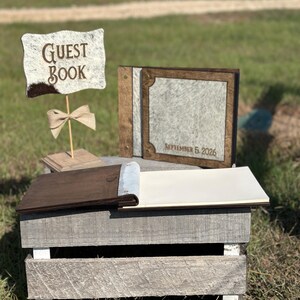 Peut inclure: Un ensemble de livre d'or comprenant un panneau avec le texte GUEST BOOK, un livre ferm&eacute; avec le texte SEPTEMBER 5, 2026, et un livre ouvert. Les objets ont des cadres en bois brun fonc&eacute; et des panneaux textur&eacute;s gris clair effet peau.