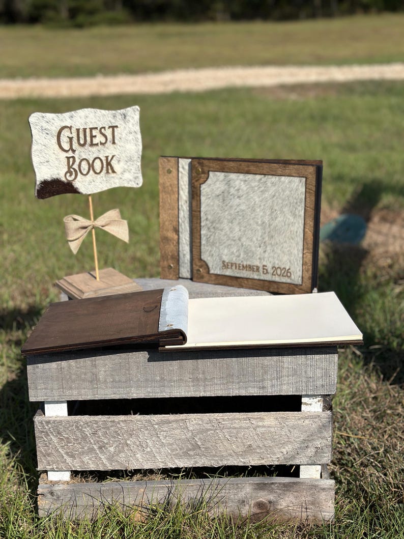 Peut inclure: Un ensemble de livre d'or comprenant un panneau avec le texte GUEST BOOK, un livre ferm&eacute; avec le texte SEPTEMBER 5, 2026, et un livre ouvert. Les objets ont des cadres en bois brun fonc&eacute; et des panneaux textur&eacute;s gris clair effet peau.