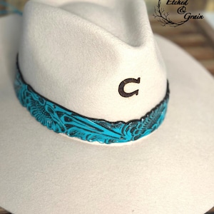 Peut inclure: Un chapeau de couleur crème arborant un symbole de fer à cheval marron foncé sur le devant et une large bande en cuir turquoise vif ornée d'un motif floral complexe. Le texte Etched & Grain est visible dans le coin supérieur droit.