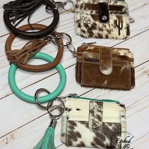 May include: Three circular key ring bracelets in dark brown, tan, and turquoise, each featuring a matching leather tassel and a cowhide card holder wallet. The wallets have snap closures and varying brown, white, and black patterns.