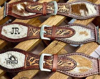 Könnte beinhalten: Vier braune Lederarmbänder für Uhren mit weiß-braunen Kuhfell-Einsätzen, silberfarbenen Metallschnallen und floralen Prägungen. Ein Armband zeigt die Initialen JR, ein weiteres ein Texas-förmiges Logo mit dem Schriftzug RED.