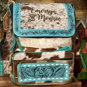 Op de afbeelding: Een bruin en wit koeienhuid rugzak met een turquoise lederen klep voorzien van reli&euml;f bloemmotieven, de naam Emersyn Monroe en een kleine koeienafbeelding. Het voorvak heeft een bijpassend turquoise lederen paneel met witte stiksels.